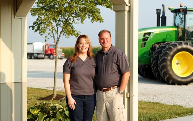 Man and woman stand together under a porch with a green tractor in the background.