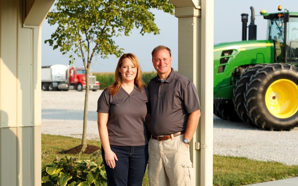 Man and woman stand together under a porch with a green tractor in the background.