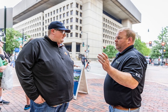 Two men talk on a sidewalk in a metropolitan area.