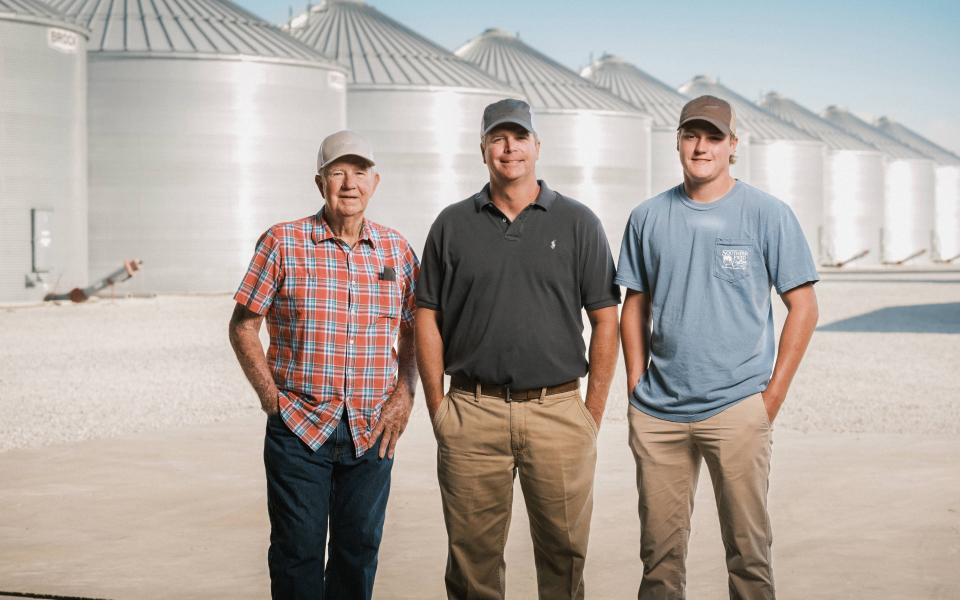 Three farmers stand in front of a line of grain bins.
