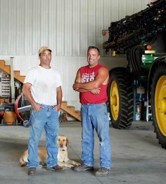 Two farmers standing in a shop next to a green sprayer.