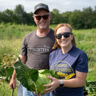 A man and woman hold a large head of cabbage in a field.