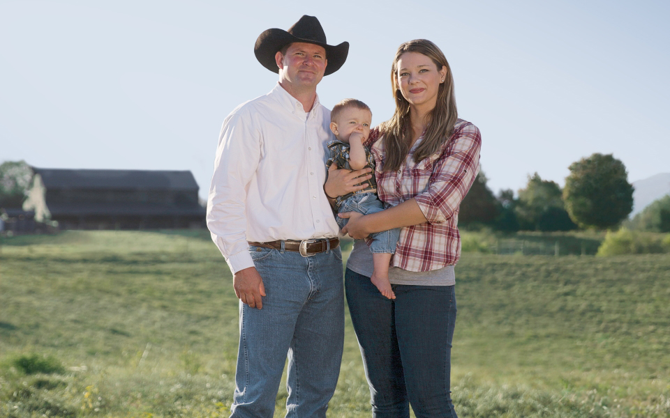 Man and woman stand with baby in a field.