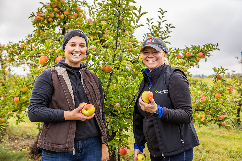 Two women holding apples freshly picked from a nearby tree.