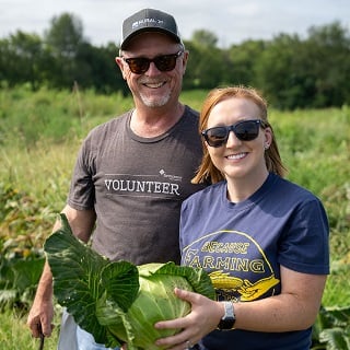 A man and woman hold a large head of cabbage in a field.
